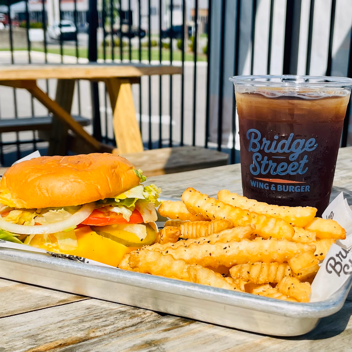 A printed clear cold cup beside a burger and crinkle fries on a metal tray, on a sunny patio table at Bridge Street Wing & Burger.
